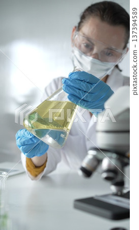 Woman scientist, wearing a lab coat, blue protective gloves, is holding an Erlenmeyer flask with yellow oily liquid in laboratory setting, portrait and vertical view. Medicine, science concept Woman scientist, wearing a lab coat, blue protective gloves, is holding an Erlenmeyer flask with yellow oily liquid in laboratory setting, portrait and vertical view. Medicine, science concept 137394589
