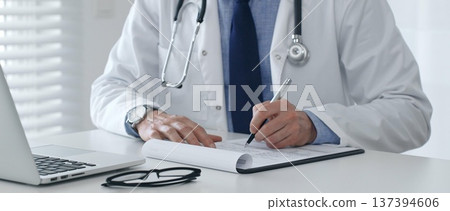 Close up of male doctor's hands filling out medical form, sitting at desk with laptop and glasses, wearing white coat and stethoscope. Medicine and health care concept 137394606