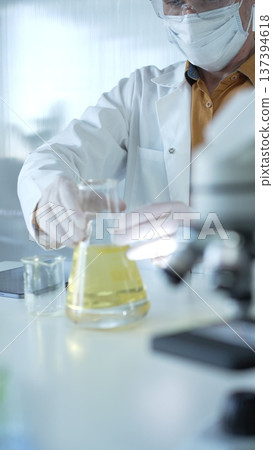 Man scientist wearing white protective gloves, mask and glasses is holding a yellow chemical solution inside an Erlenmeyer flask in a laboratory, vertical portrait. Science and medicine concept Man scientist wearing white protective gloves, mask and glasses is holding a yellow chemical solution inside an Erlenmeyer flask in a laboratory, vertical portrait. Science and medicine concept 137394618