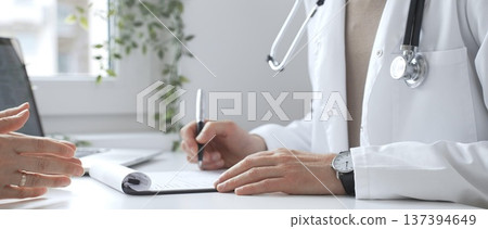 Doctor listening to patient health concerns and writing a prescription during a consultation in a bright, modern clinic. Medicine and health care concept Doctor listening to patient health concerns and writing a prescription during a consultation in a bright, modern clinic. Medicine and health care concept 137394649
