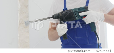 Close up of unrecognizable male construction worker wearing blue overalls and protective white gloves, is demolishing white wall with rotary hammer drill, generating dust, horizontal view. Renovation Close up of unrecognizable male construction worker wearing blue overalls and protective white gloves, is demolishing white wall with rotary hammer drill, generating dust, horizontal view. Renovation 137394651