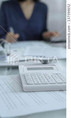 Businesswoman wearing blue dress is working with laptop, calculator and documents in a professional office setting. Low lighting, close-up vertical view. Business people, audit concept Businesswoman wearing blue dress is working with laptop, calculator and documents in a professional office setting. Low lighting, close-up vertical view. Business people, audit concept 137394652