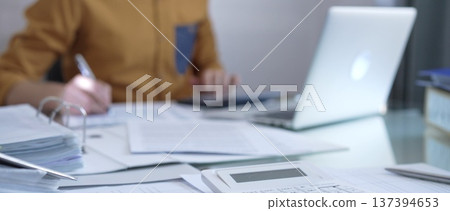 Magnifying glass, calculator, pen and documents are lying on accountant's desk. Businessman or accountant, wearing yellow shirt, is working with documents on the background. Business people, audit Magnifying glass, calculator, pen and documents are lying on accountant's desk. Businessman or accountant, wearing yellow shirt, is working with documents on the background. Business people, audit 137394653
