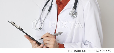 Close up of female doctor wearing stethoscope and white coat over brick red blouse is standing and writing on a clipboard, representing medical expertise and patient care. Medicine concept 137394660