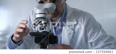 Male scientist wearing lab coat and protective glasses using microscope doing research in modern laboratory setup, portrait view. Low key lighting photo. Science and Medicine concept 137394661