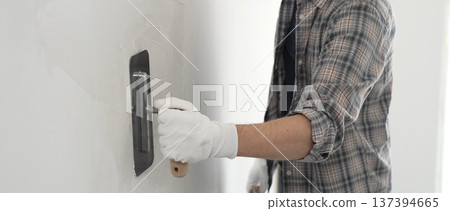 Unknown male construction worker wearing gloves and plaid shirt is using finishing trowel to plaster a white wall, applying a layer of plaster to create a smooth surface during renovation project Unknown male construction worker wearing gloves and plaid shirt is using finishing trowel to plaster a white wall, applying a layer of plaster to create a smooth surface during renovation project 137394665