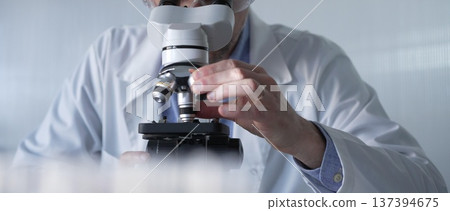 Male scientist wearing lab coat and protective glasses using microscope doing research in modern laboratory setup, portrait view. Low key lighting photo. Science and Medicine concept Male scientist wearing lab coat and protective glasses using microscope doing research in modern laboratory setup, portrait view. Low key lighting photo. Science and Medicine concept 137394675