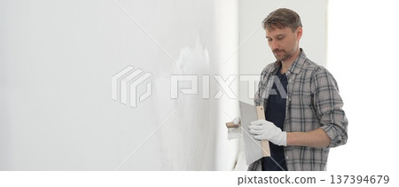 Male construction worker wearing protective gloves and plaid shirt, spreading plaster smoothly across wall surface using professional drywall taping knife during renovation project. Portrait view Male construction worker wearing protective gloves and plaid shirt, spreading plaster smoothly across wall surface using professional drywall taping knife during renovation project. Portrait view 137394679