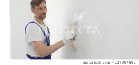 Male construction worker wearing protective gloves and blue construction coveralls, spreading plaster smoothly across wall using professional drywall taping knife during renovation project. Portrait Male construction worker wearing protective gloves and blue construction coveralls, spreading plaster smoothly across wall using professional drywall taping knife during renovation project. Portrait 137394693