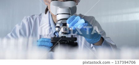 Woman scientist wearing lab coat, white protective gloves and glasses, is using microscope in modern laboratory setup, portrait view. Low key lighting photo. Science and Medicine Woman scientist wearing lab coat, white protective gloves and glasses, is using microscope in modern laboratory setup, portrait view. Low key lighting photo. Science and Medicine 137394706