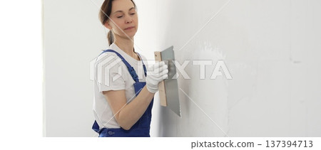 Woman construction worker wearing protective gloves and blue construction coveralls, spreading plaster smoothly across wall using professional drywall taping knife during renovation project. Portrait Woman construction worker wearing protective gloves and blue construction coveralls, spreading plaster smoothly across wall using professional drywall taping knife during renovation project. Portrait 137394713