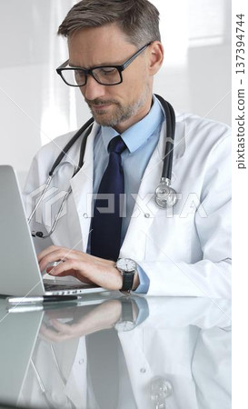 Male doctor wearing spectacles, thinking about healthcare diagnosis while using a laptop in a clinic office. Medicine and health care Male doctor wearing spectacles, thinking about healthcare diagnosis while using a laptop in a clinic office. Medicine and health care 137394744