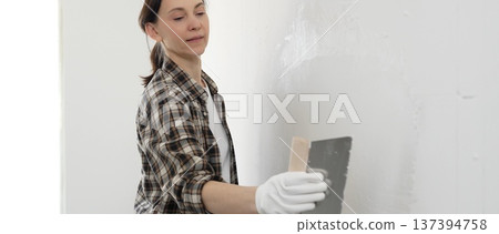 Woman construction professional wearing checkered shirt is smoothing white wall surface, applying putty with professional spatula technique, showcasing skilled interior renovation work 137394758