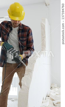 Male construction worker wearing red checkered shirt, yellow hard hat and protective gloves, is demolishing white wall with rotary hammer drill, generating dust, medium long shot. Renovation project 137394760