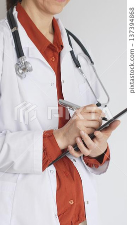 Unknown woman doctor wearing stethoscope and white coat over brick red blouse is standing and writing on a clipboard, representing medical expertise and patient care. Medicine and health care concept 137394868