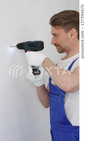 Man construction worker in a blue jumpsuit, safety goggles, and work gloves is screwing a screw into the wall using a cordless screwdriver, carrying out a careful home renovation. Vertical portrait Man construction worker in a blue jumpsuit, safety goggles, and work gloves is screwing a screw into the wall using a cordless screwdriver, carrying out a careful home renovation. Vertical portrait 137394870
