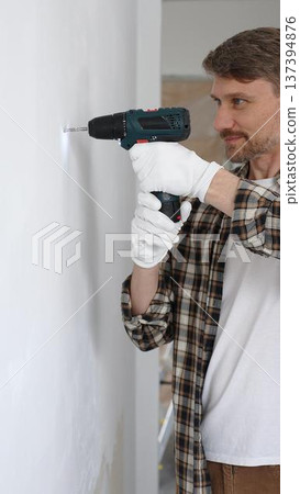 Middle aged man wearing checked shirt and white gloves, is drilling hole in a wall with cordless drill during home renovation work. Portrait view Middle aged man wearing checked shirt and white gloves, is drilling hole in a wall with cordless drill during home renovation work. Portrait view 137394876
