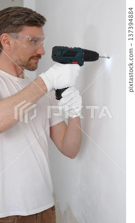 Middle aged man worker wearing white t-shirt and protective gloves, is drilling hole in a wall with cordless drill during home renovation work. Vertical portrait view 137394884