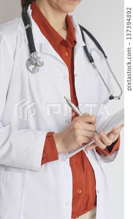 Close up of woman doctor wearing stethoscope and white coat over brick red blouse is standing and writing on a clipboard, representing medical expertise and patient care. Medicine concept Close up of woman doctor wearing stethoscope and white coat over brick red blouse is standing and writing on a clipboard, representing medical expertise and patient care. Medicine concept 137394892