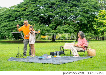 Asian senior man stand in the back of little girl who play song with violin and senior woman also sit on chair with glad during family relax in public park together with day light. Asian senior man stand in the back of little girl who play song with violin and senior woman also sit on chair with glad during family relax in public park together with day light. 137395456