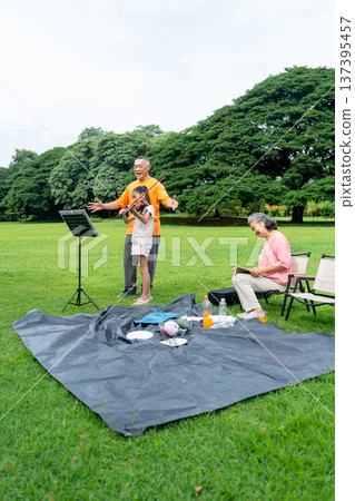 Vertical image of Asian senior man stand in the back of little girl who play song with violin and senior woman also sit on chair with glad during family relax in public park together with day light. 137395457