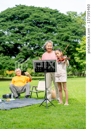 Vertical image of Asian senior woman stand in the back of little girl who play song with violin and senior man also sit on chair with glad during family relax in public park together with day light. Vertical image of Asian senior woman stand in the back of little girl who play song with violin and senior man also sit on chair with glad during family relax in public park together with day light. 137395460