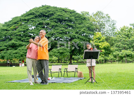 Asian senior man and woman enjoy to dancing while the girl play song with violin in the public park during relax with family. Asian senior man and woman enjoy to dancing while the girl play song with violin in the public park during relax with family. 137395461