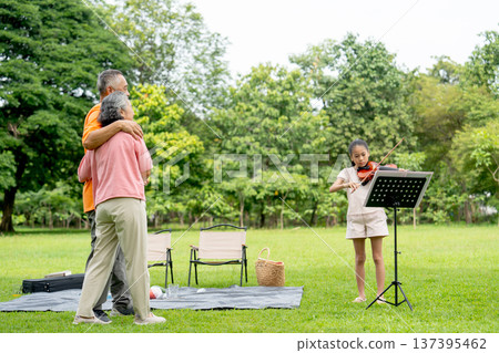 Asian senior man and woman enjoy to dancing while the girl play song with violin in the public park during relax with family. 137395462