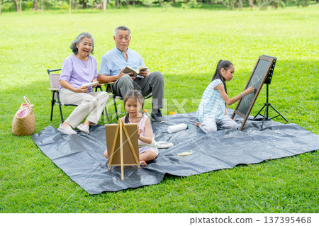 Senior man read the book and sit beside of senior woman while two little girls enjoy to draw or paint the picture in public park with day light. Senior man read the book and sit beside of senior woman while two little girls enjoy to draw or paint the picture in public park with day light. 137395468