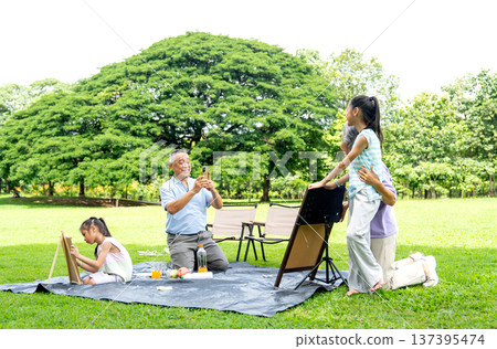 Asian senior man use mobile phone to take a photo of senior woman hug with the girl and hold the drawing during relax with family in public park together with day light. Asian senior man use mobile phone to take a photo of senior woman hug with the girl and hold the drawing during relax with family in public park together with day light. 137395474