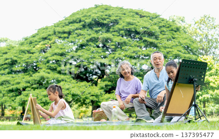 Lower view of Asian senior man use mobile phone to take a picture during the girl draw or paint and senior woman also look at her work in public park and big tree in the background. 137395477
