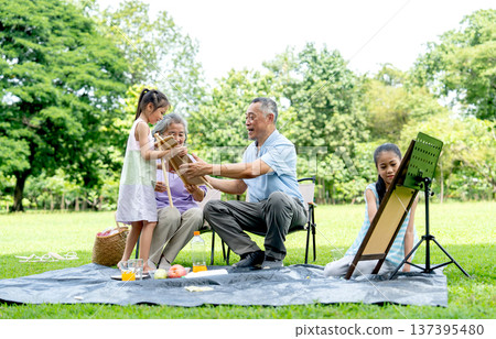 Asian senior man and woman express glad and look at the drawing of one girl while the other girl concentrate to draw or paint in public park with day light. 137395480