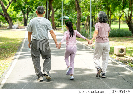 Back of Asian girl hold hands of senior man and woman and walk along the road in public park together for exercise and get good health for the family. Back of Asian girl hold hands of senior man and woman and walk along the road in public park together for exercise and get good health for the family. 137395489