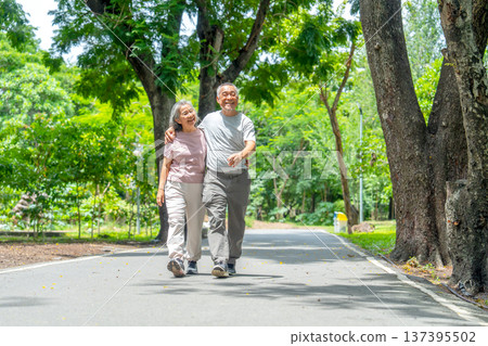 Wide shot of Asian senior man put his arm around senior woman and walk along the road in public park with happiness during exercise and relax together. Wide shot of Asian senior man put his arm around senior woman and walk along the road in public park with happiness during exercise and relax together. 137395502