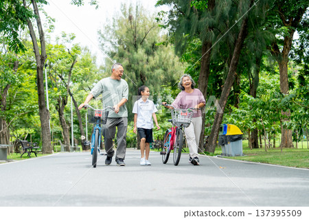 Wide shot of Asian grandmother and grandfather leading the bicycles with the girl who walk on the road together in the public park with day light. Wide shot of Asian grandmother and grandfather leading the bicycles with the girl who walk on the road together in the public park with day light. 137395509