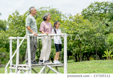 Asian grandfather and grandmother stand on tall wooden stand together with grandchild and they look forwad and smile in public park. Asian grandfather and grandmother stand on tall wooden stand together with grandchild and they look forwad and smile in public park. 137395512