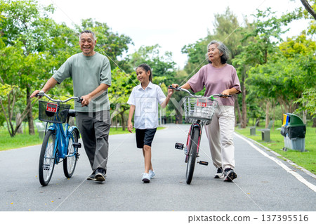 Front view of senior man and woman leading the bicycles with the girl who walk on the road together in the public park with day light. 137395516