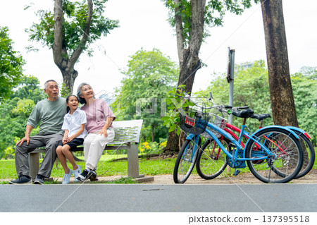 Senior man and woman sit on garden bench with the girl and look to their left side and two bicycle set beside of them in the public park with day light. Senior man and woman sit on garden bench with the girl and look to their left side and two bicycle set beside of them in the public park with day light. 137395518