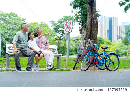 Asian grandfather and grandmother or senior man and woman look at the direction that the girl point to when they sit on garden bench in the public park with happiness. Asian grandfather and grandmother or senior man and woman look at the direction that the girl point to when they sit on garden bench in the public park with happiness. 137395519