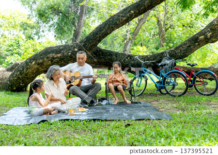 Front view of Asian grandfather and grandmother enjoy and relax with two girls in public park with eating fruit and play music and sing a song together. 137395521