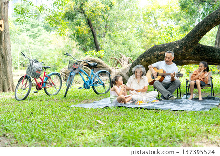 Asian grandfather and grandmother enjoy and relax with two girls in public park with eating fruit and play music and sing a song together. Asian grandfather and grandmother enjoy and relax with two girls in public park with eating fruit and play music and sing a song together. 137395524
