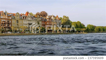 Buildings line the shore of a lake with trees beside the water on a sunny day Buildings line the shore of a lake with trees beside the water on a sunny day 137395609
