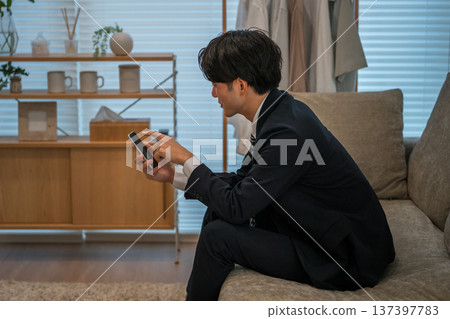 一位身穿西裝的亞洲商人夜晚坐在沙發上，看著他的智慧型手機。 137397783