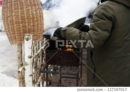 Vendor in winter attire serving steaming soup outdoors beside wicker basket and fence 137397917