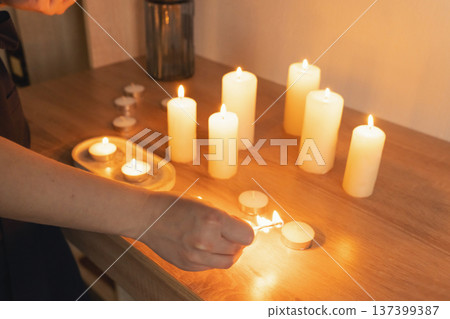 Hand lighting small candle with match, surrounded by multiple glowing pillar candles creating warm, relaxing ambiance, atmospheric glow Hand lighting small candle with match, surrounded by multiple glowing pillar candles creating warm, relaxing ambiance, atmospheric glow 137399387