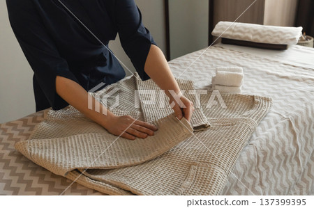 On massage table, a woman's hands fold a clean waffle robe next to neatly folded white towels. 137399395