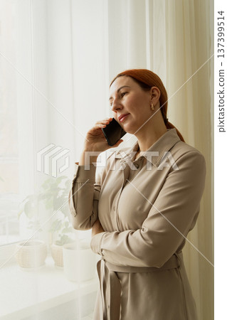 Woman is having a business conversation, standing by the window with her smartphone, feeling thoughtful and socializing while working from home 137399514