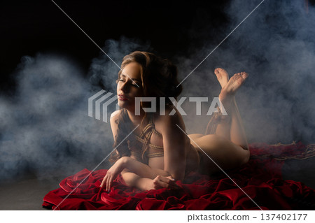 Serene woman tied in shibari ropes lying on red fabric in a smoky, dark setting Serene woman tied in shibari ropes lying on red fabric in a smoky, dark setting 137402177