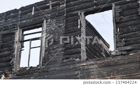Charred ruins of a burnt wooden log house with empty window frames against an overcast sky. Charred ruins of a burnt wooden log house with empty window frames against an overcast sky. 137404224