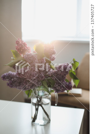 Spring flowers in home interior. Blooming lilac in vase. Sunny still life with opened book by window. Bouquet of beautiful lilac flowers and book. Purple, blue, pink, white bouquet. Copy space 137405727